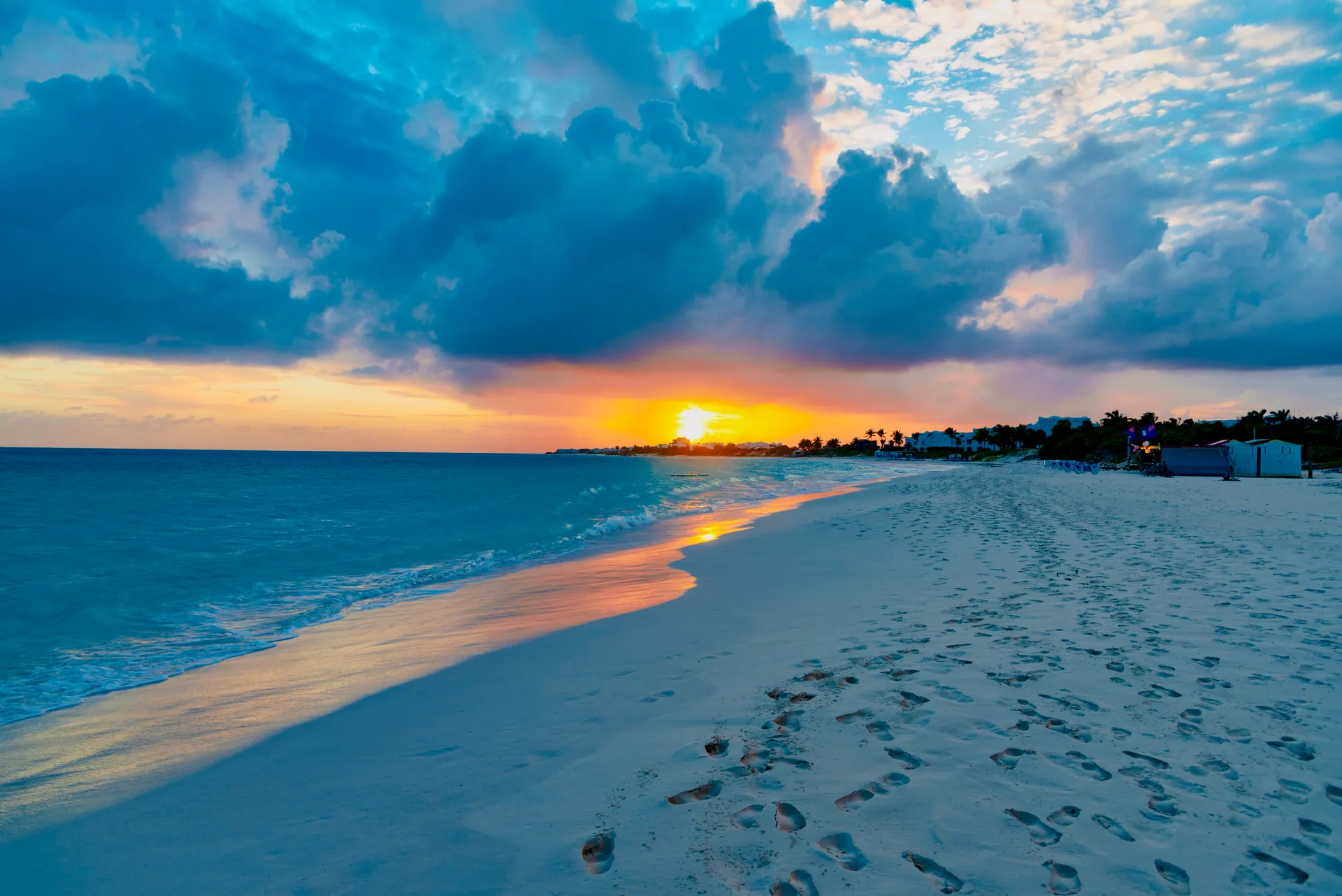 Sunset over turquoise water, with footprints across white sand and palms along the shoreline